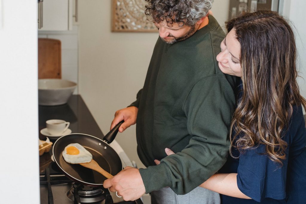 Side view of crop adult man frying egg for breakfast while wife gently hugging in light contemporary kitchen