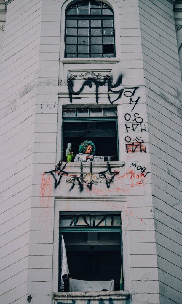 From below of positive female with colorful curly hair standing near window of residential building with vandalized facade